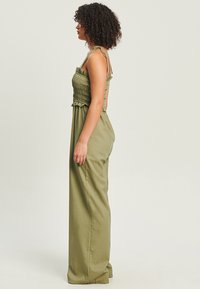 Combinaison vert olive avec un corsage smocké et des pantalons longs et décontractés. Comprend des bretelles ajustables et un tissu léger et texturé.
