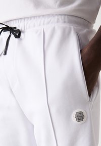 Pantalon de jogging blanc avec cordon noir, doté d'une poche sur le côté et d'un patch logo rond. Texture lisse avec des plis personnalisés.