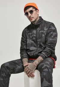 Svart camo-jakke og bukser, glat materiale, lynlås lukning, orange hue, runde solbriller, synlige tatoveringer på hænderne. Enkel baggrund.