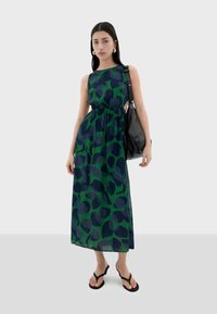 Abito maxi in verde e blu navy con motivo astratto a foglie, realizzato in tessuto leggero. Presenta una vita aderente e aperture laterali. La modella tiene in mano una borsa nera.