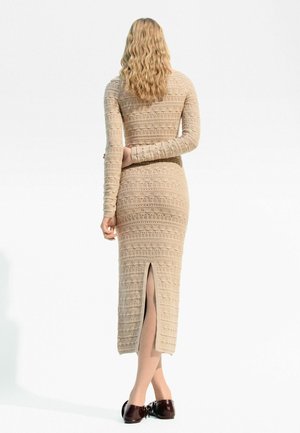 Vestito beige lavorato a maglia con maniche lunghe e spacco posteriore, caratterizzato da motivi testurizzati. Indossato con scarpe marrone scuro, visto da dietro.