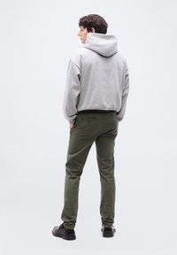 Νέος άνδρας που στέκεται με την πλάτη γυρισμένη, φοράει ανοιχτό γκρι φούτερ με κουκούλα, παντελόνι ελιάς και μαύρα παπούτσια σε απλό λευκό φόντο.