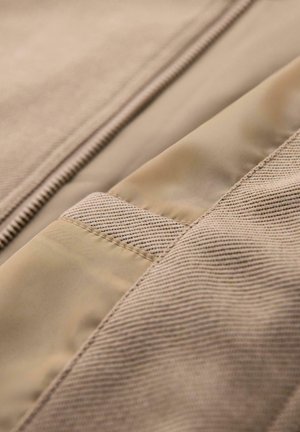 Beige stof med en blanding af glatte og strukturerede materialer, med detaljeret syning og en skjult lynlås langs kanten.