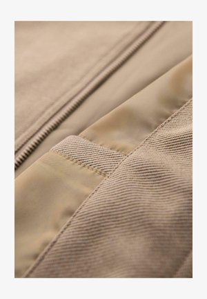 Beige stof med en blanding af glatte og strukturerede materialer, med detaljeret syning og en skjult lynlås langs kanten.