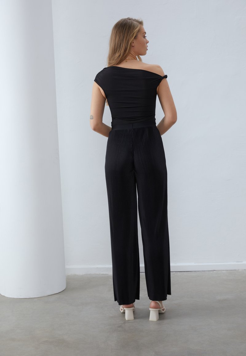 Top nero scollato con design aderente, abbinato a pantaloni neri a vita alta, plissettati e a gamba larga, e sandali con tacco a blocco beige.