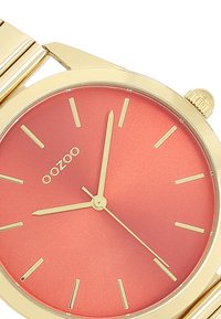 Montre-bracelet dorée avec un boîtier rond, un cadran rouge texturé, des index horaires dorés et des aiguilles fines. Nom de marque "oozoo" affiché.