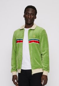 Zöld velúr cipzáras dzseki, bézs gallérral, piros és kék csíkos díszítéssel, valamint egy arany logóval a bal mellen.