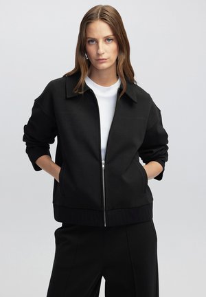 Chaqueta negra con cremallera y cuello, que presenta un diseño estructurado, puños acanalados, bolsillos laterales y una textura suave. Llevada sobre una camiseta blanca.