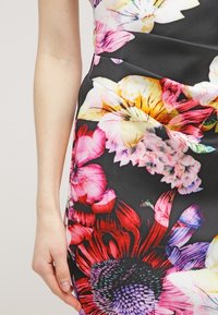 Gros plan sur la main d'une personne reposant à côté d'une robe ajustée avec de grands motifs floraux colorés sur fond noir.
