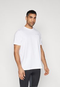 Camiseta blanca de manga corta hecha de tela ligera, con un logo sutil en el pecho y un ajuste relajado, combinada con leggings negros.