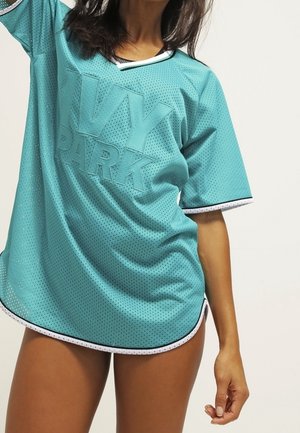 Kobieta w oversize'owej turkusowej siateczkowej sportowej koszulce z nadrukiem "IVY PARK" na przodzie, stojąca na tle jednolitego jasnego tła.