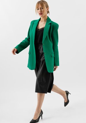 Blazer verde con revers a punta, chiusura con un bottone e due tasche frontali, abbinato a una maglia nera e culotte in pelle. Tacchi neri.