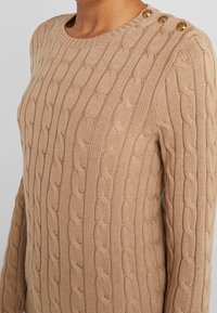 Suéter beige de punto con un patrón de trenzas, cuello redondo y tres botones decorativos dorados en el hombro. Tejido texturizado.