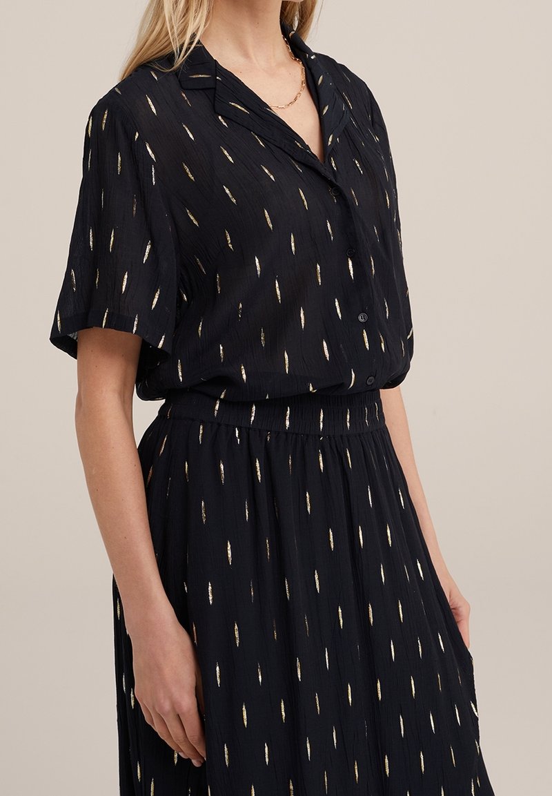 Mujer con camisa negra abotonada y falda con patrones de rayas metálicas doradas y un collar de cadena dorada, de pie frente a un fondo beige.