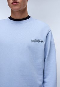 Lys blå sweatshirt med lange ermer, rund hals og en liten svart logo på brystet. Glatt tekstur, uformell passform.
