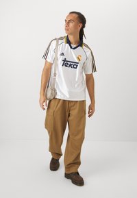 Maillot de soccer blanc adidas avec des accents bleus et jaunes, pantalon cargo beige et bottes marron. Comprend un petit sac à bandoulière.
