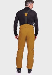 Brown waterproof snow overalls with adjustable black suspenders and a smooth texture, paired with black ski boots and a fitted black top.