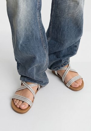 Pieds portant des sandales plates à brides tressées argentées associées à un jean bleu délavé et ample sur un fond uni.