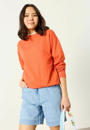 Mladá žena oblečená v oranžovom tričku s dlhým rukávom a svetlomodrých rifľových šortkách, držiaca farebný remienok tašky s kvetinovým vzorom.