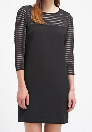 Frau in einem schwarzen, knielangen Kleid mit halbtransparenten, horizontal gestreiften Ärmeln und Passe, die vor einem einfarbigen Hintergrund steht.