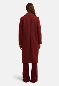 Femme debout de dos, portant un long manteau bordeaux sur un pantalon bordeaux ample avec des rayures latérales, sur un fond clair uni.