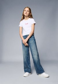 Bijela majica kratkih rukava s crvenim logom, u kombinaciji s svijetloplavim širokim trapericama i bijelim tenisicama, na sivom pozadini.