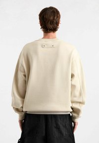 Felpa oversized beige con collo rotondo, polsini a coste e dettaglio a toppa sul retro. Texture liscia, materiale morbido, design informale.