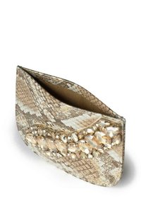 Kopertówka w beżowo-kremowym wzorze przypominającym skórę węża, o zaokrąglonym kształcie, ozdobiona przezroczystymi kamieniami na przodzie. Teksturowane wykończenie.