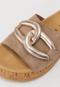 Beige suede slide sandal with a shiny gold chain accent, cork-like sole, and soft texture. Rounded shape with an open toe design.