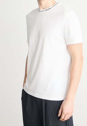 Mand iført en ensfarvet hvid kortærmet T-shirt med "BOSS" på kraven og sorte bukser med tynde striber, overkrop synlig, hånd hvilende ved siden.