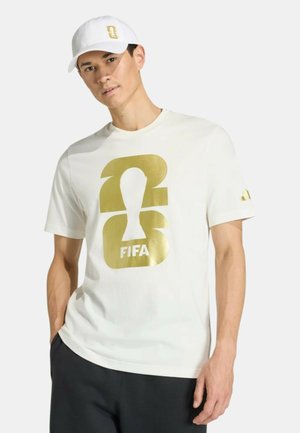Uomo che indossa un cappellino bianco e una maglietta con un grande logo FIFA oro, in piedi con una posa rilassata contro uno sfondo semplice.