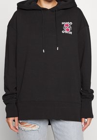 Sudadera con capucha negra de corte holgado, que presenta un logotipo floral en rosa y blanco en el pecho. Tiene cordones y puños acanalados.