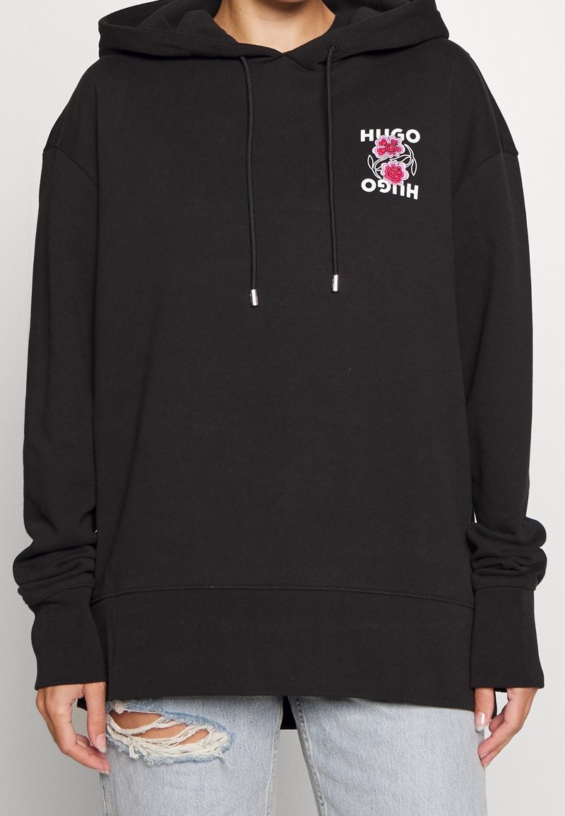 Sudadera con capucha negra de corte holgado, que presenta un logotipo floral en rosa y blanco en el pecho. Tiene cordones y puños acanalados.