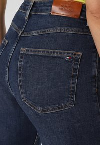 Jeans in denim blu scuro con un'etichetta in pelle marrone con "Tommy Hilfiger" e un piccolo emblemma della bandiera sulla tasca posteriore. Vestibilità classica.