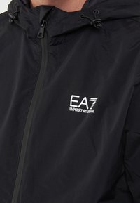 Black windbreaker jacket with a zip closure and adjustable drawstring hood. Features a white EA7 logo on the chest. Smooth, lightweight fabric.