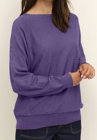 Lila stickad tröja med båtringning, ribbade ärmslut och fåll. Mjuk textur och avslappnad passform. Bärs med mörka jeans.