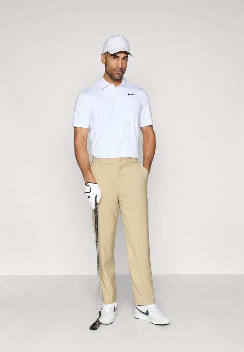 Golfeur masculin en polo blanc avec un logo noir, pantalon beige, baskets blanches, tenant un club de golf, portant une casquette blanche et un gant.