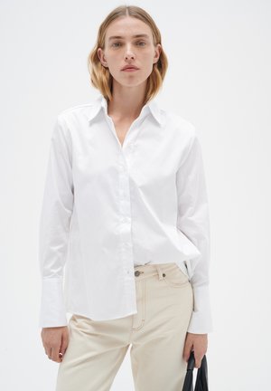 Chemise blanche à boutons en coton avec col, manches longues et coupe décontractée, associée à un pantalon beige clair et un sac noir.