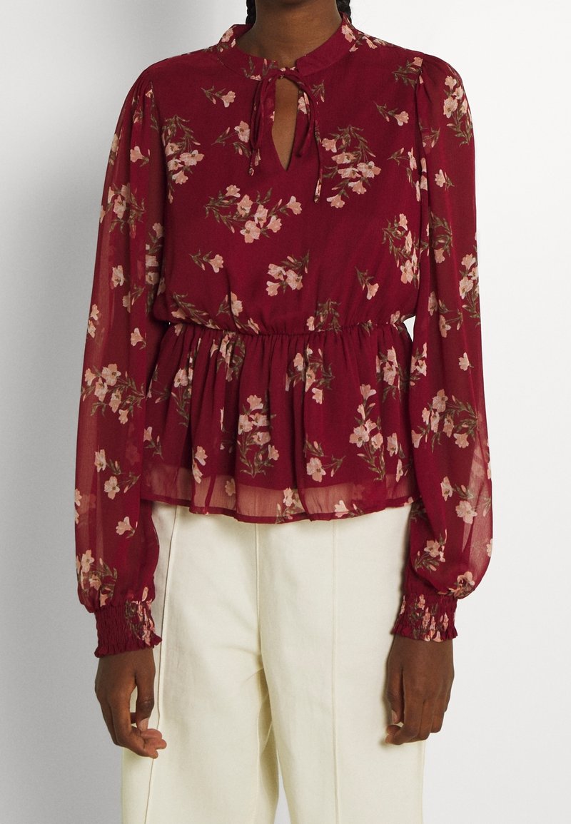 Femme portant un chemisier rouge à imprimé floral avec de longues manches transparentes, un col clé et une taille froncée, ainsi qu'un pantalon blanc taille haute.