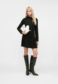 Svart, langermet kjole med innsvingt midje og vidt skjørt, kombinert med knelangt, blankt svart støvler og en hvit håndveske.