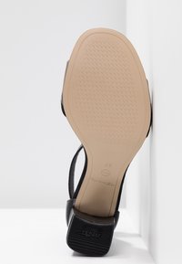 Semelle d'une sandale noire avec une semelle beige texturée, taille 37, et un talon noir antidérapant, positionnée contre des murs blancs.