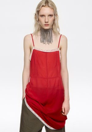 Młoda kobieta o blond włosach nosi czerwoną sukienkę na ramiączkach na zielonych spodniach oraz duży srebrny naszyjnik z frędzlami na tle białego tła.