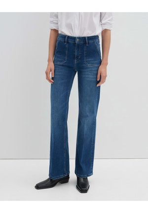 Osoba ubrana w ciemnoniebieskie jeansy o prostych nogawkach, białą koszulę z podwiniętymi rękawami oraz czarne botki o kwadratowym nosku, stojąca na jednolitym tle.