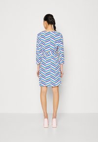 Kurzärmeliges Kleid im mehrfarbigen Zickzackmuster (blau, orange, weiß) mit einem Taillengürtel; dazu hellrosa Blockabsatzschuhe.