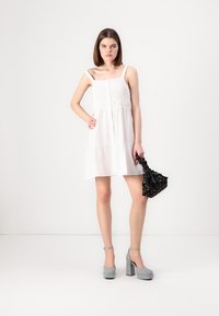 Λευκό αμάνικο φόρεμα με δαντελένιες λεπτομέρειες, tiered σχέδιο και κουμπιά. Το μοντέλο κρατάει μια μαύρη τσάντα με λουλούδια και φοράει γκρίζα παπούτσια με χοντρό τακούνι.