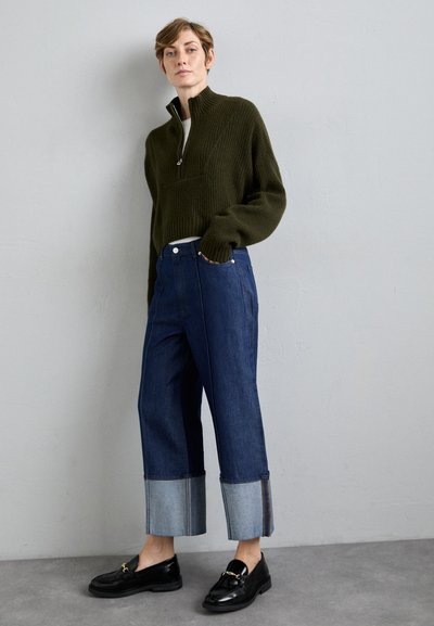 Person wearing dark green zip-up sweater, wide-leg cuffed blue jeans, and black loafers, standing against a plain light gray wall.