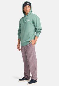 Felpa verde menta con tasca frontale, cordini neri e logo bianco. Abbinata a pantaloni in corduroy lavanda dal taglio ampio e sneakers scure.