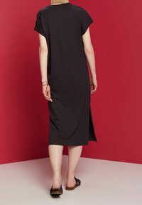 Robe midi en coton noir avec des manches courtes, des fentes latérales, une coupe décontractée et un design simple, associée à des sandales plates sur un fond rouge.