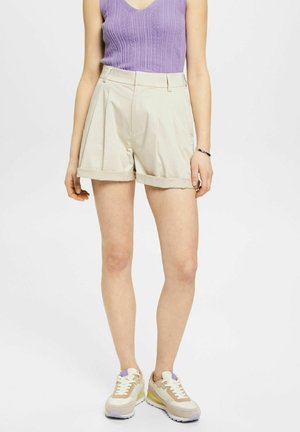 Femme portant un short beige plissé à ourlet roulé, un haut sans manches en laine violet et des baskets beiges avec des lacets blancs et des accents jaunes.