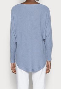 Light blue long-sleeve knit sweater worn by person standing with back to camera, paired with white pants against plain background.